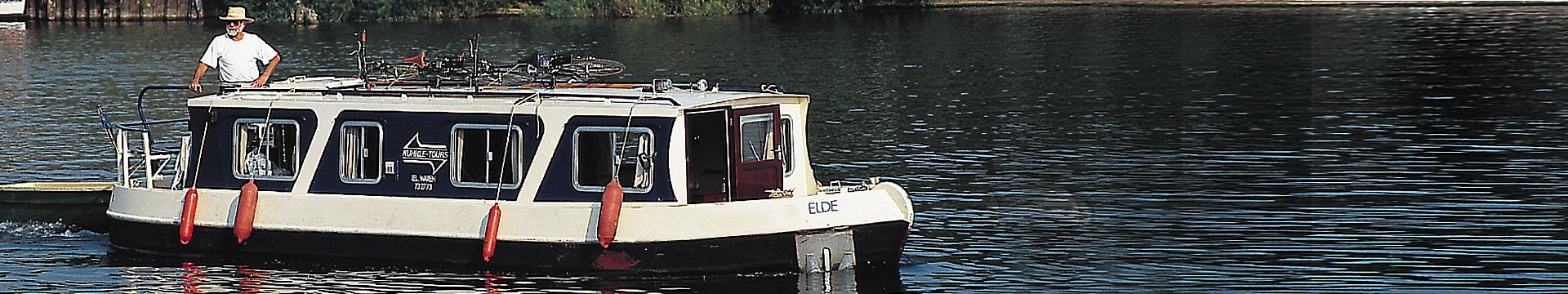 Ein älterer Herr mit Bart, der an einen echten Kapitän erinnert, fährt mit einem Hausboot im Stil eines englischen Narrowbootes über einen See.