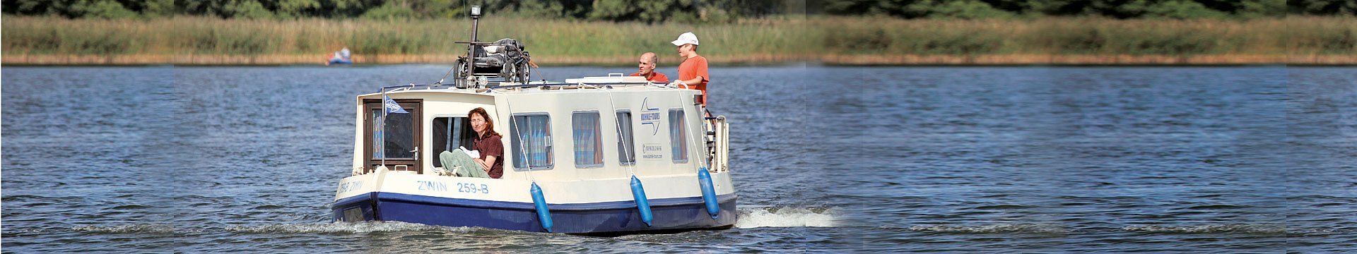 Eine kleine Familie fährt auf einem Hausboot im Stil eines englischn Naroowbootes auf einem See. Die Mutter sitzt vorn, Vater und und Sohn stehen hinten.